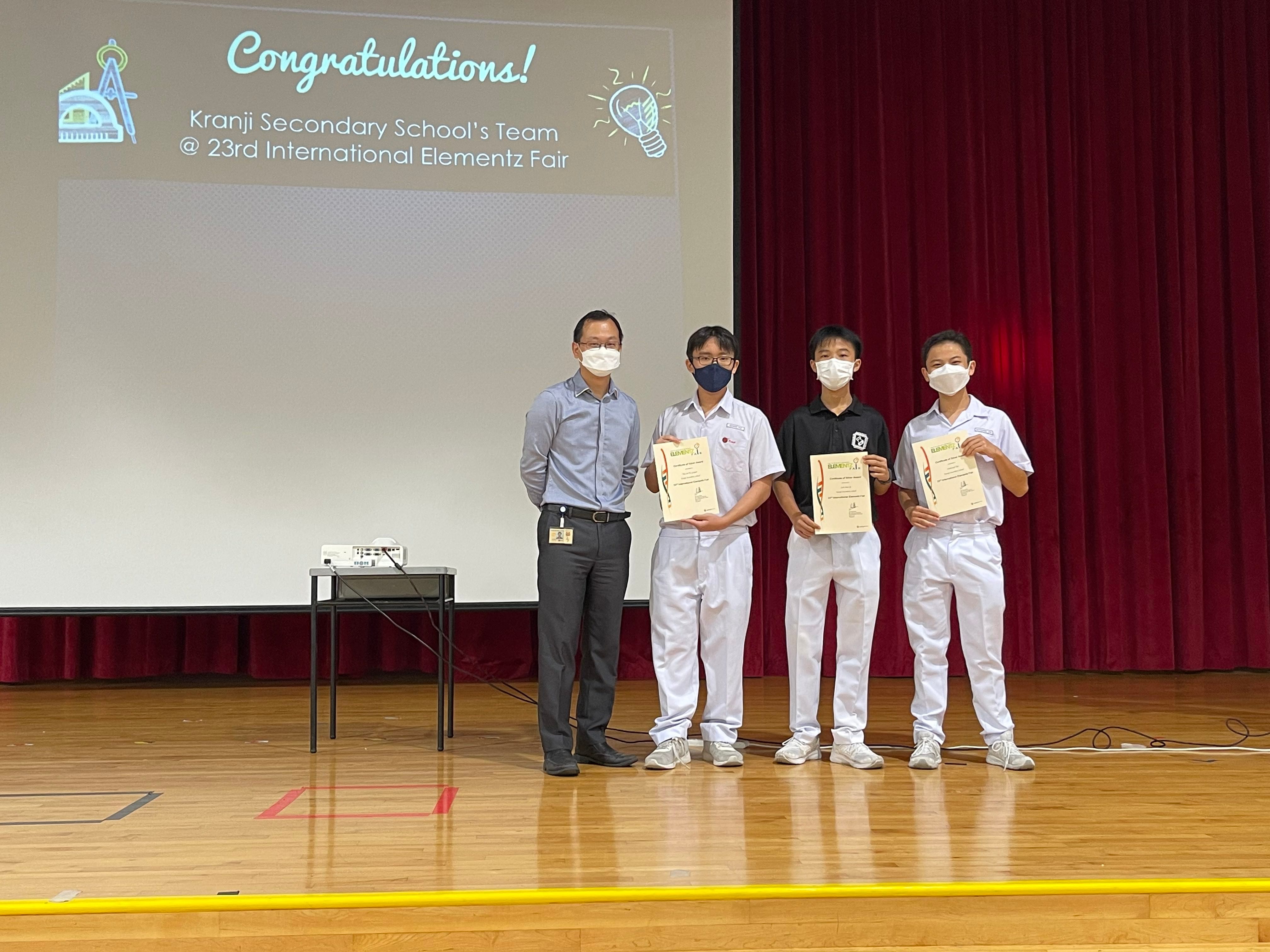 Four masked people stand on a school stage holding certificates, with a congratulations slide for Kranji Secondary School visible behind them.